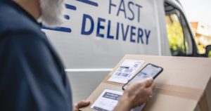 Close-up of mature bearded independent delivery expert holding package and scanning label details with smart phone.