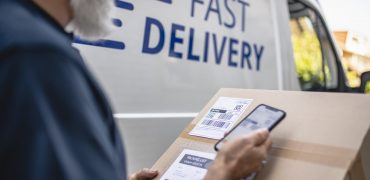 Close-up of mature bearded independent delivery expert holding package and scanning label details with smart phone.
