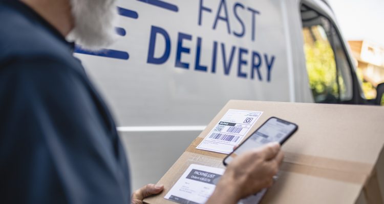 Close-up of mature bearded independent delivery expert holding package and scanning label details with smart phone.