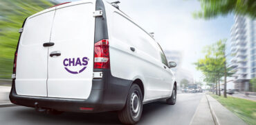 Rear view of a white CHAS van driving in a city. Motion blurred city background with buildings and trees.
